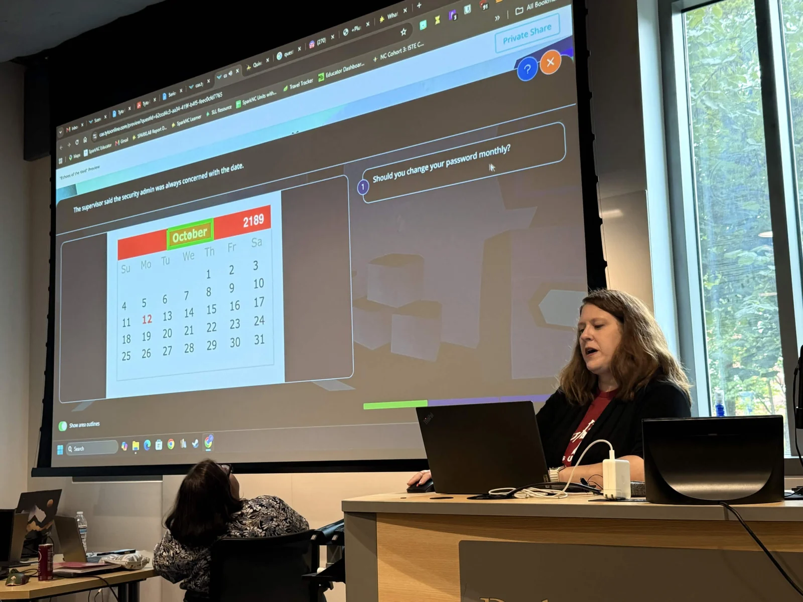 An attendee presenting her finished quest from the lectern, with an in-game calendar puzzle and the dialogue question “Should you change your password monthly?” projected behind her.