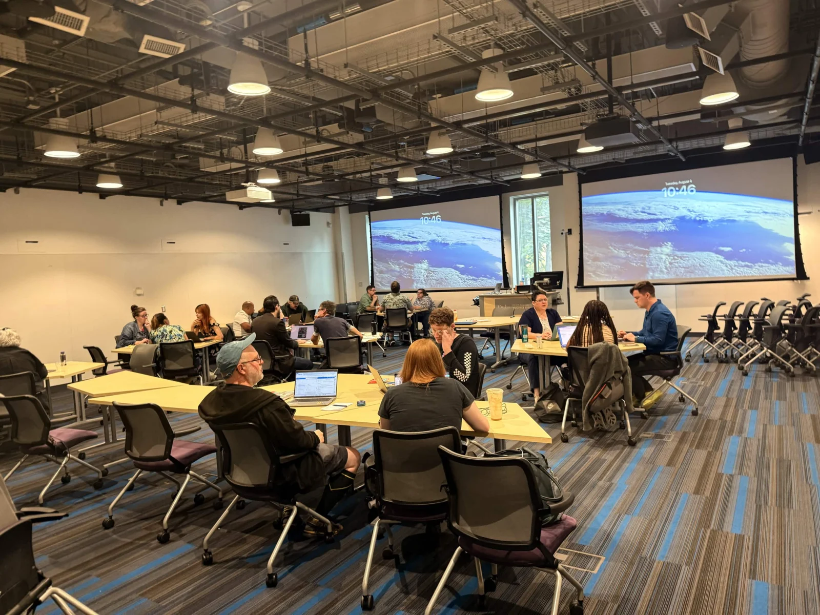 Attendees spread across tables in twos and threes, laptops open, working while a planet-from-orbit scene sits on the projection screens behind them.