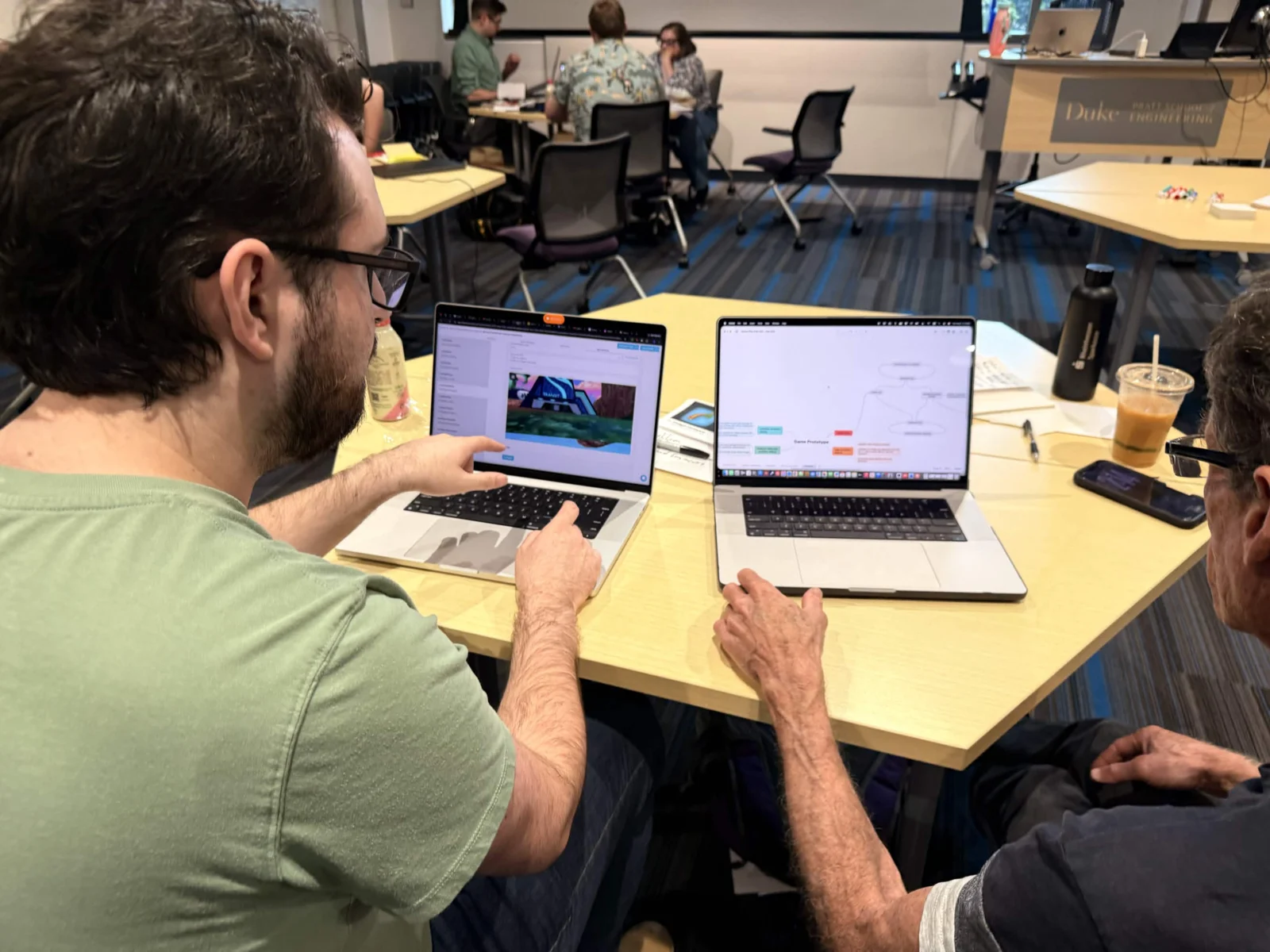 Two attendees working side by side, one pointing at a 3D game scene on his laptop while the other has a branching flow diagram open on hers.