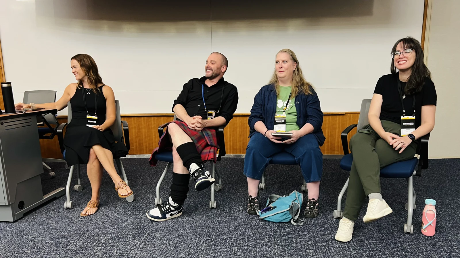 Four panelists seated in a row at the front of a conference room, all laughing mid-discussion, conference lanyards on.